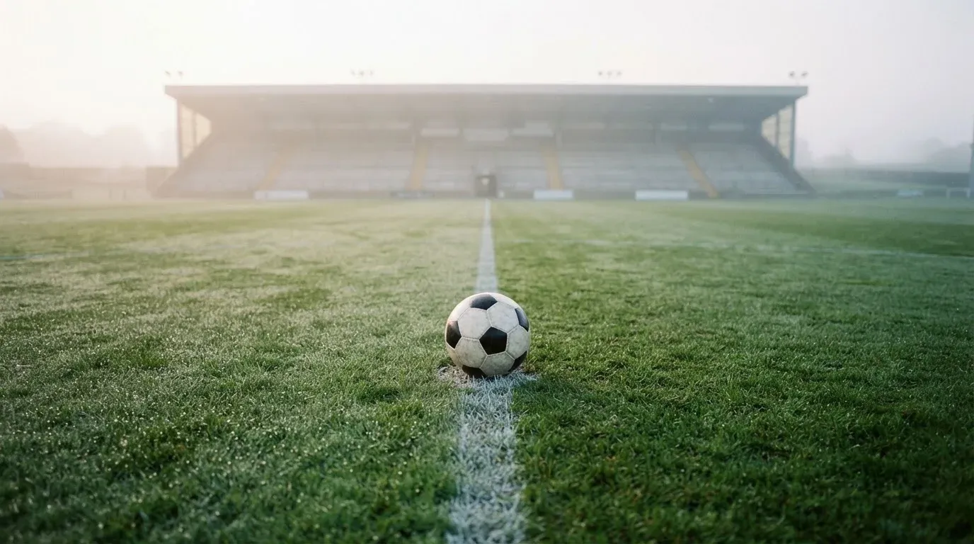 Ein einzelner Fussball liegt auf dem Anstosspunkt eines leeren Rasenplatzes am frühen Morgen, Tau glitzert auf dem Gras, die Tribüne liegt im Dunst.