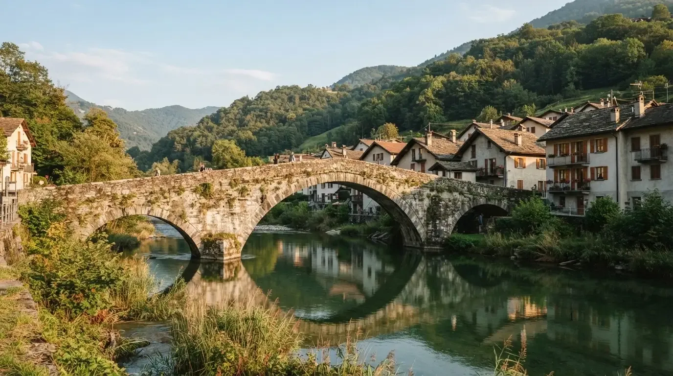 Eine alte Steinbrücke über einen ruhigen Fluss, dahinter Häuser mit Ziegeldächern und bewaldete Hügel.