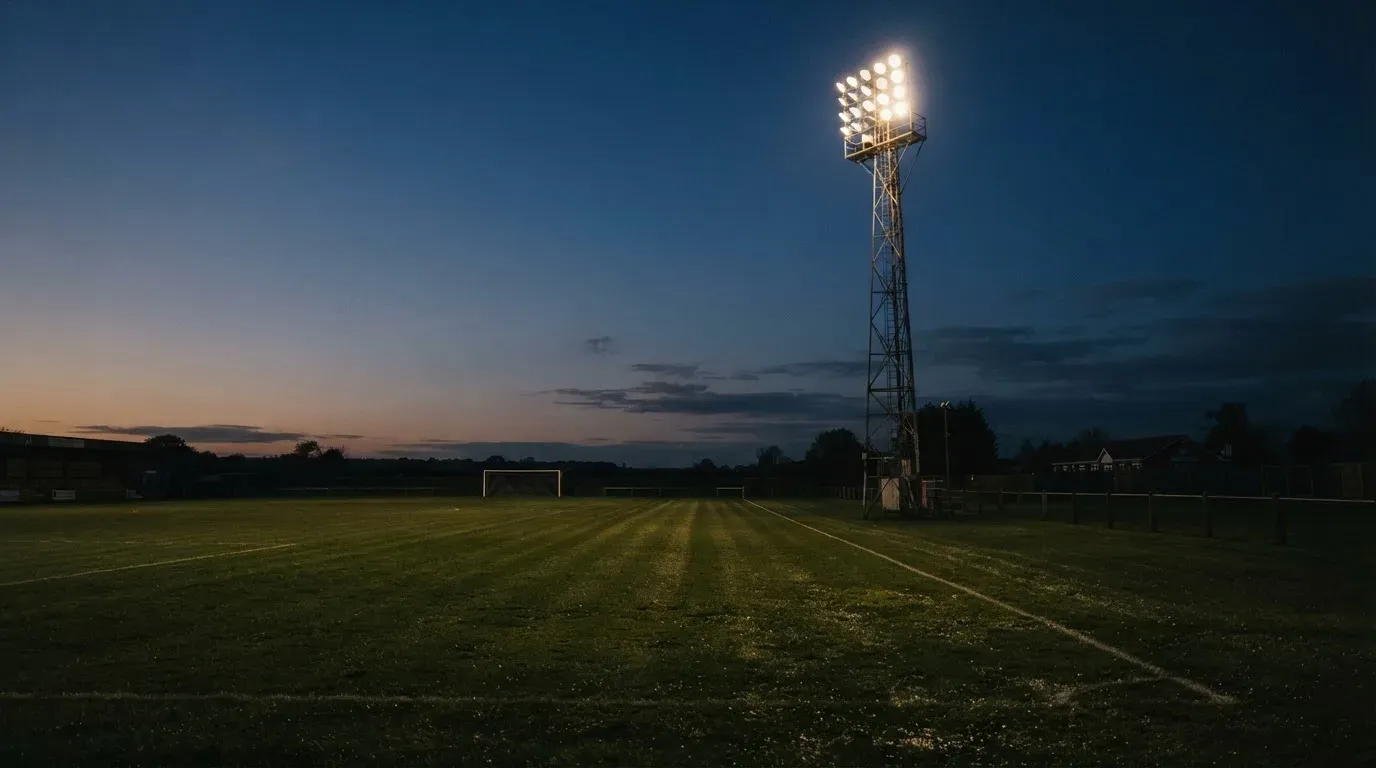 Ein Flutlichtmast ragt in den Abendhimmel, die Rasenfläche darunter liegt leer und dunkelgrün da.