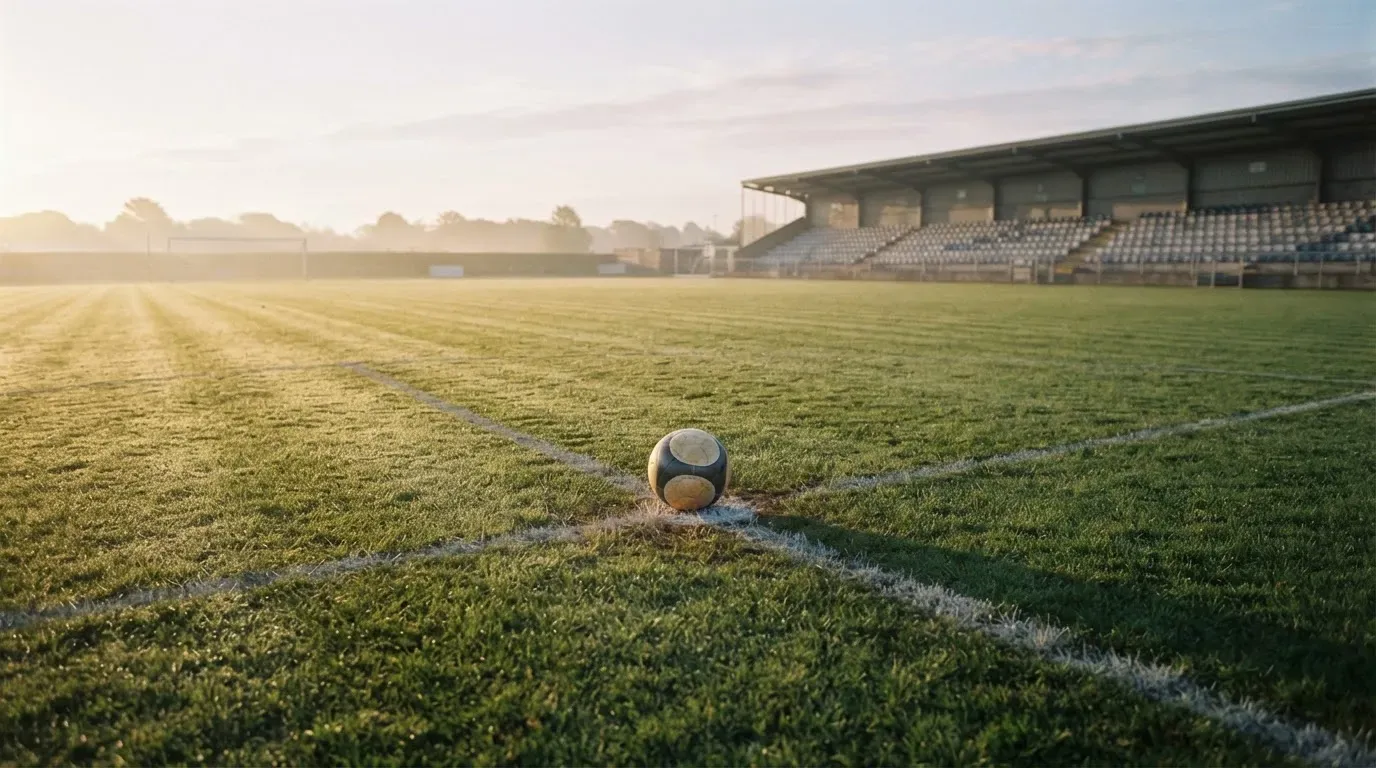 Ein Fussball liegt auf dem Anstosspunkt eines leeren Rasenplatzes im frühen Morgenlicht, dahinter erhebt sich eine menschenleere Tribüne.