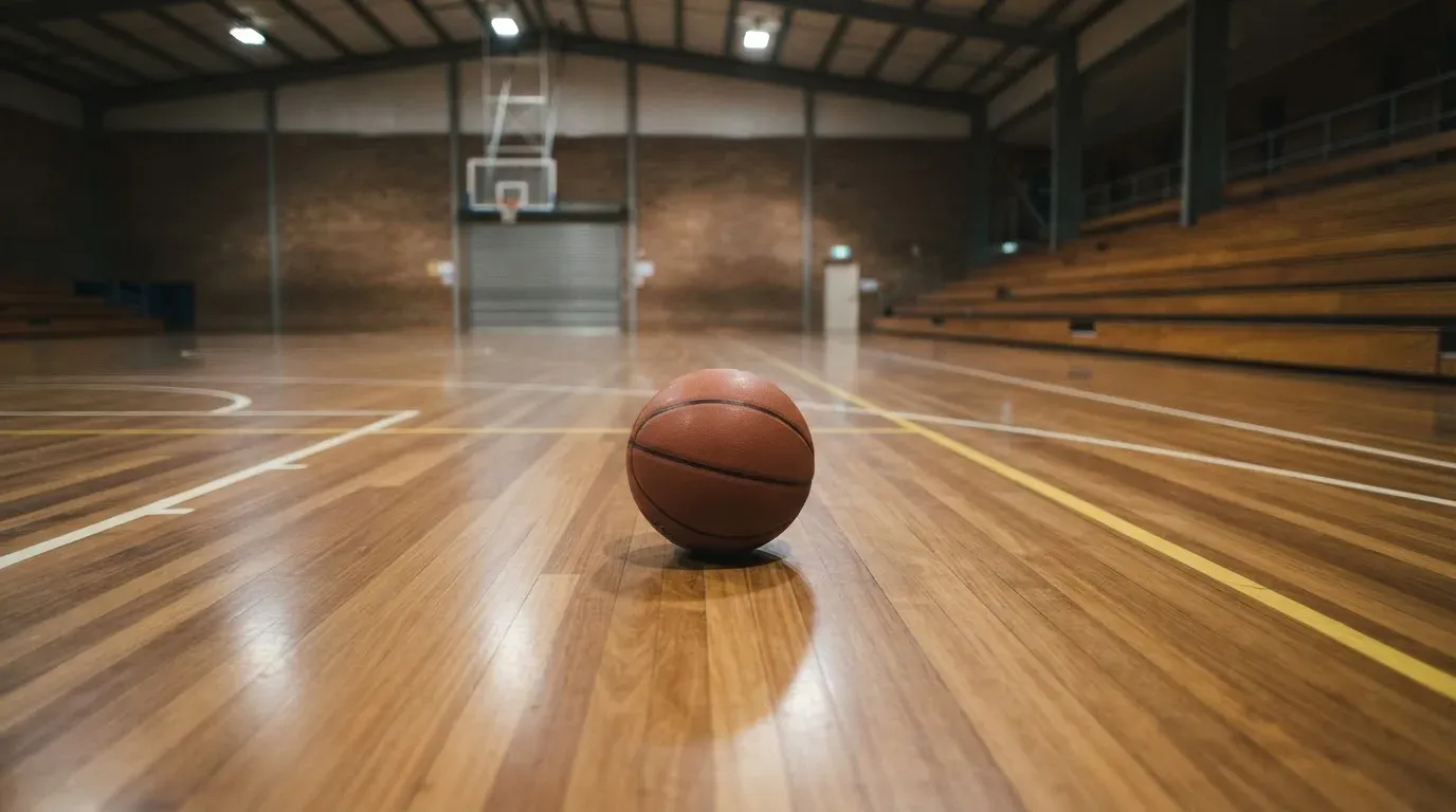 Ein Basketball liegt auf dem Parkett einer leeren Halle, von den Deckenlampen fällt gleichmässiges Licht auf den Boden.
