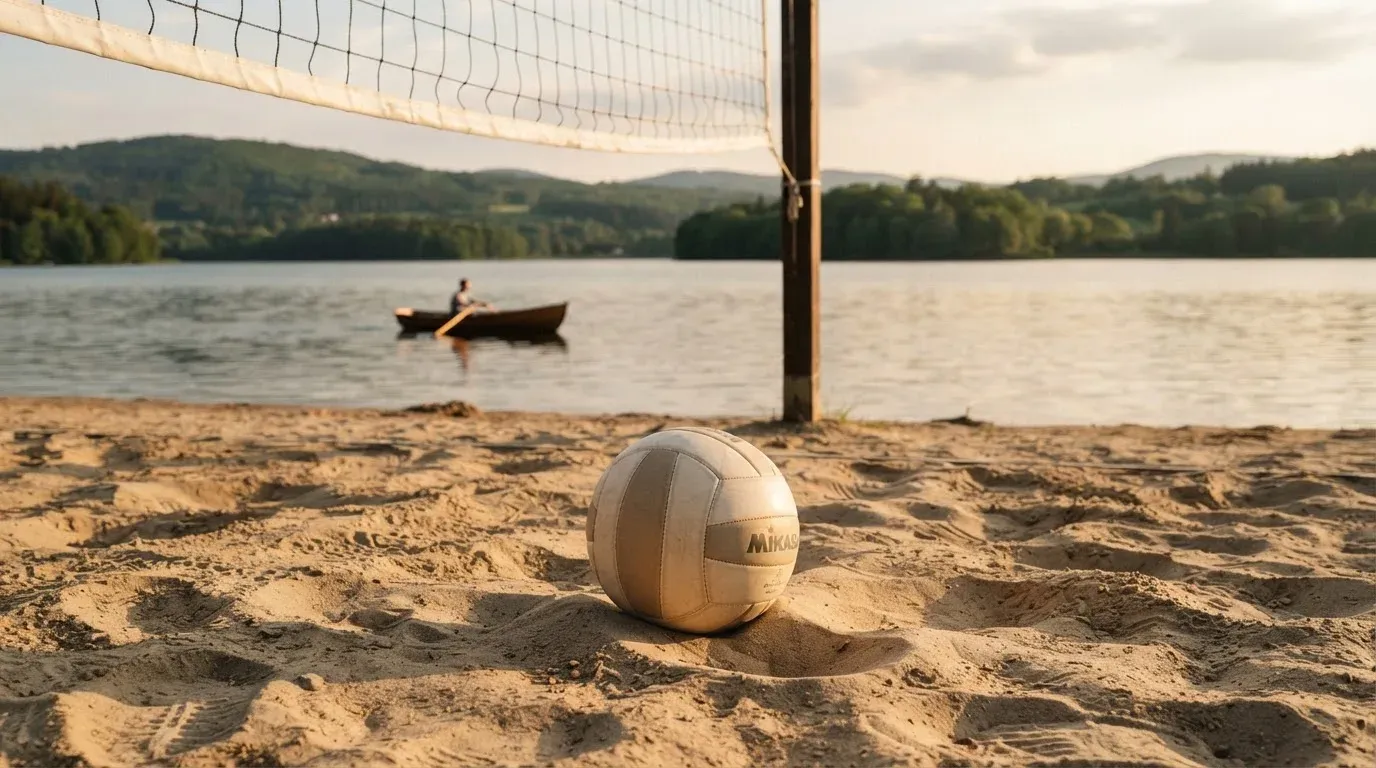 Ein Volleyball liegt im Sand eines Spielfelds am Seeufer, im Hintergrund gleitet ein Boot über das Wasser.