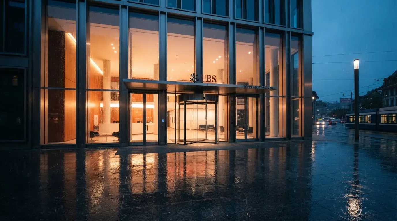 Eine Fassade aus Glas und Stahl am Eingang eines Bürogebäudes im Regen, auf dem nassen Boden spiegelt sich das Licht.