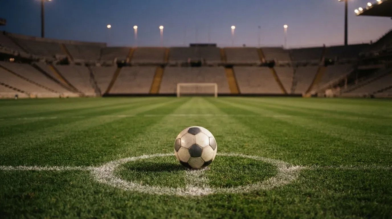 Ein Fussball liegt auf dem Mittelkreis eines leeren Rasenplatzes, dahinter ragen leere Tribünenreihen in den Abendhimmel.