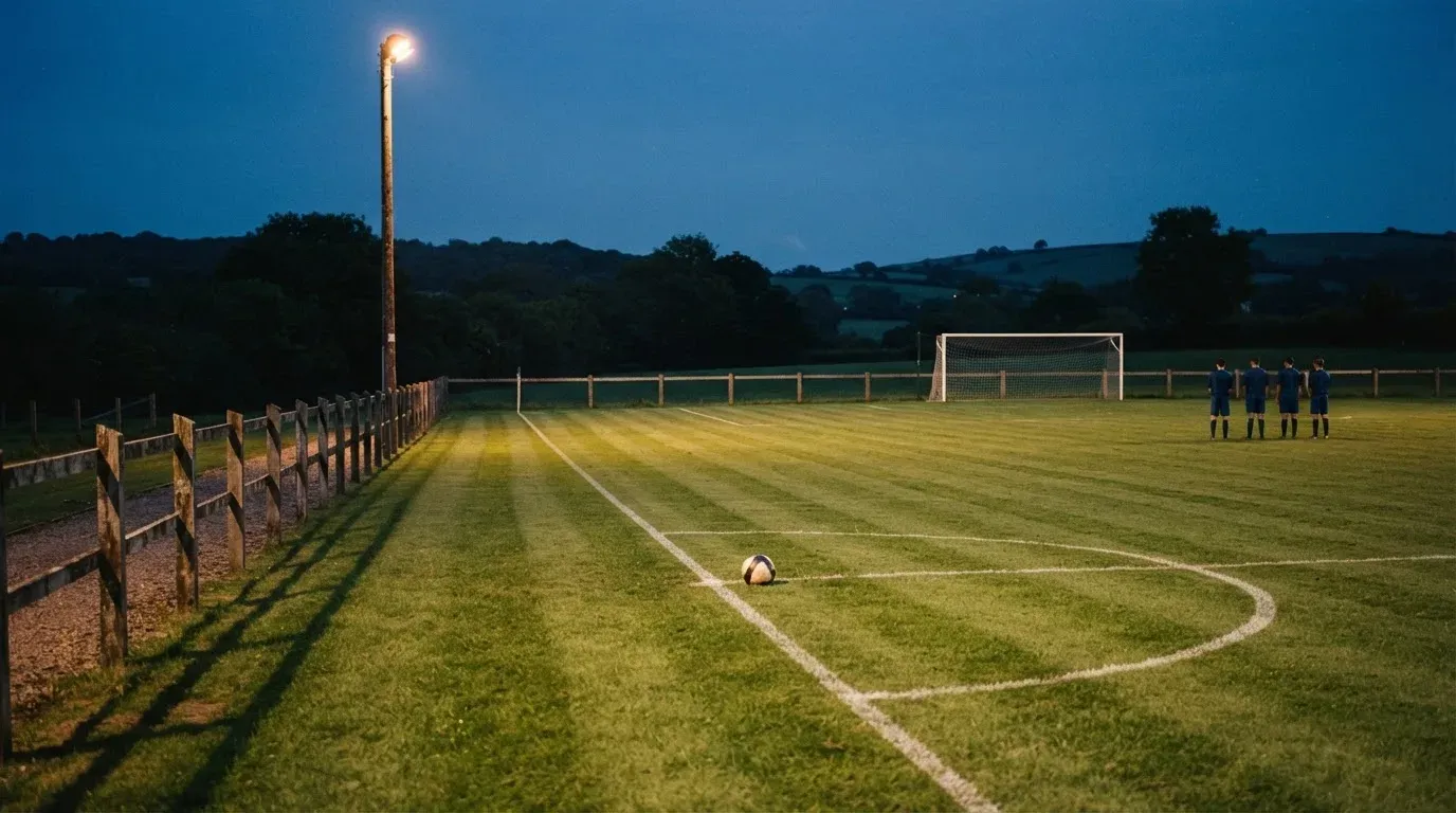 Ein kleiner Dorf-Fussballplatz liegt in der Dämmerung unter einem hohen Flutlichtmast, am Spielfeldrand steht ein Holzzaun.