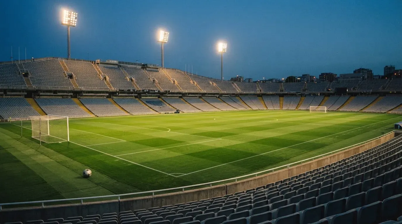 Leere Tribünenränge eines Fussballstadions in der Abenddämmerung, über dem Rasen leuchten die Flutlichter.