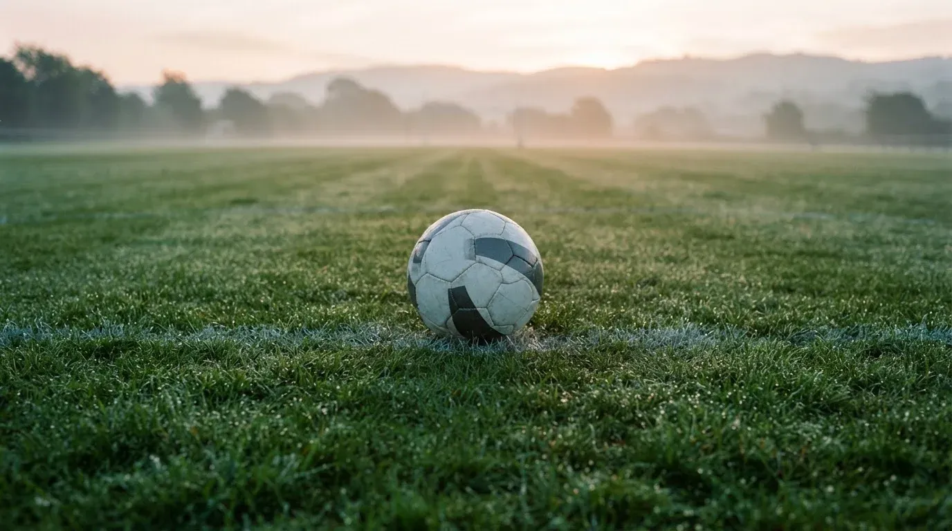 Ein Fussball liegt am frühen Morgen auf dem Anstosspunkt eines taunassen Rasens, im Hintergrund erheben sich Hügel im Dunst.