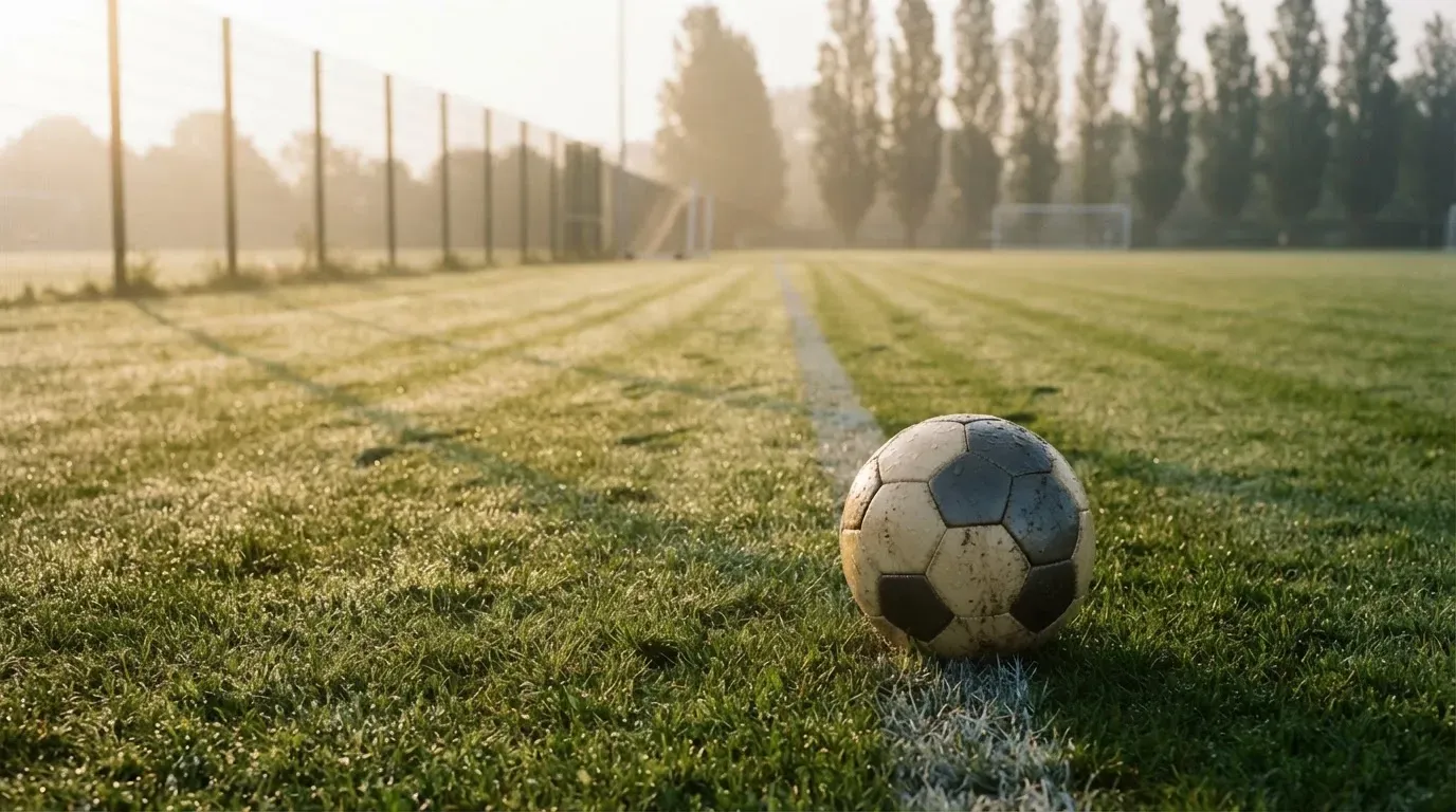 Ein Fussball ruht an der Seitenlinie im taufeuchten Gras, die ersten Sonnenstrahlen fallen schräg über den Platz.