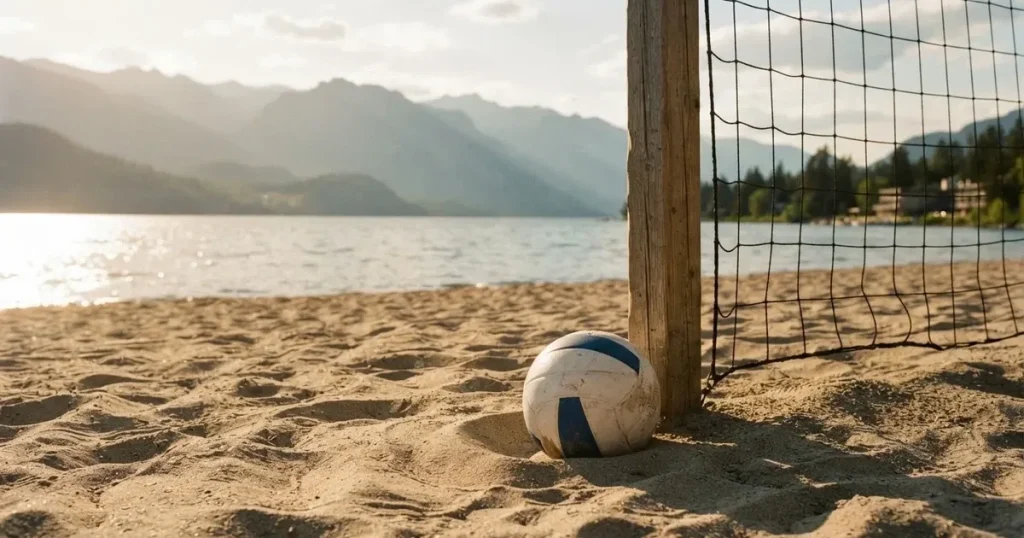 Ein Volleyball liegt am Fuss des Netzes auf Sand, im Hintergrund glitzert ein See und ein Bergzug steht im Dunst.