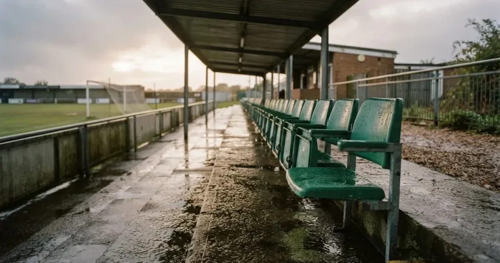 Eine Reihe leerer Sitzschalen auf der Tribüne eines kleinen Stadions, auf den Stufen glänzt Regenwasser.
