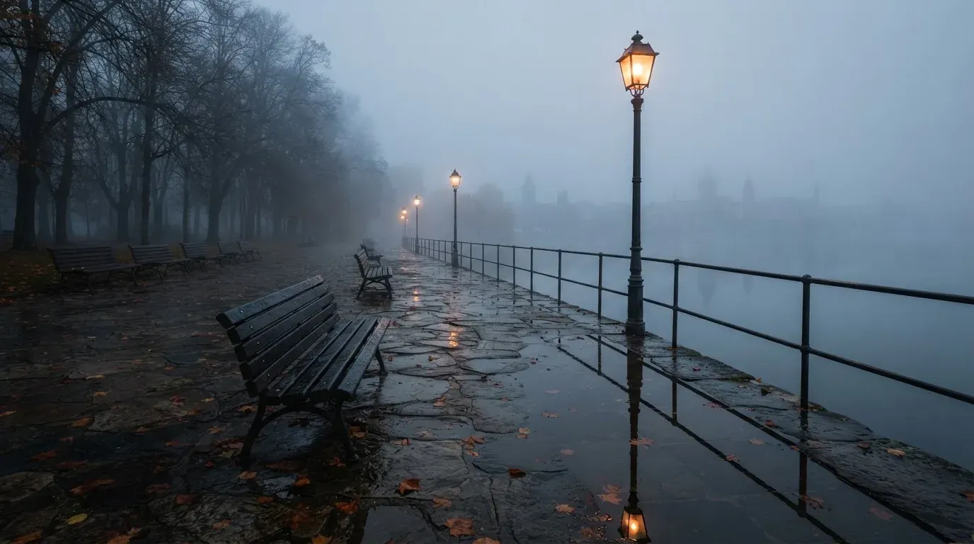 Eine Seepromenade mit leeren Bänken an einem nebligen Morgen, im Wasser spiegeln sich die Lichter der Uferlaternen.