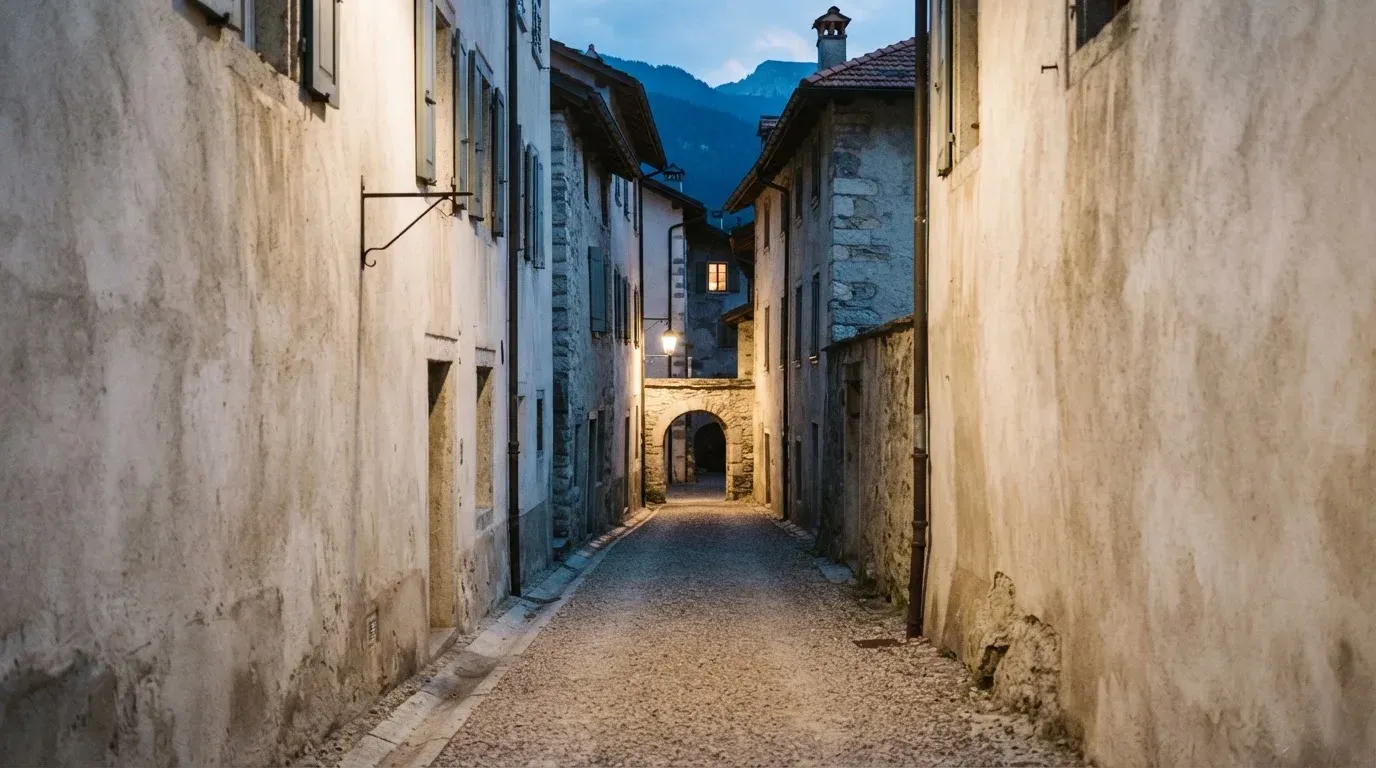 Ein schmaler Kiesweg führt zwischen zwei hellen Mauern durch eine Schweizer Altstadt, die Laternen sind noch nicht angezündet.