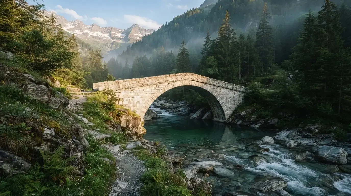Eine Steinbogenbrücke überspannt einen Bergbach in den Alpen im Morgenlicht, an den Hängen stehen dunkle Nadelbäume.