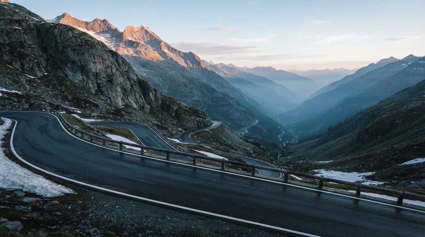 Ein Bergpass in den Schweizer Alpen mit leerer Strasse und Schneeresten am Rand im ersten Frühlicht.