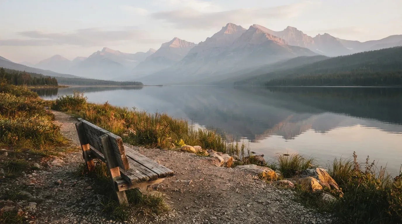 Eine Holzbank an einem Seeufer mit Blick auf eine Bergkette im Morgendunst, das Wasser ist spiegelglatt.