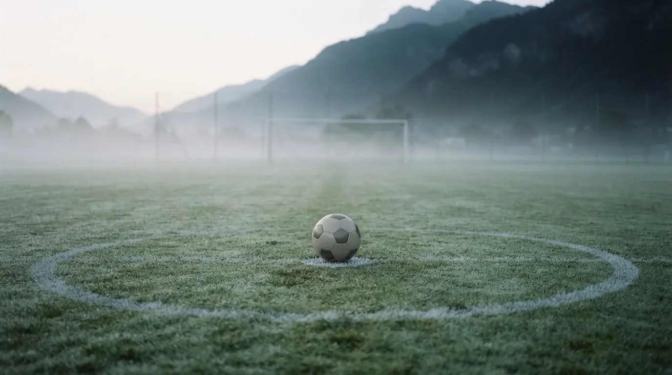 Ein einzelner Fussball auf dem Mittelpunkt eines leeren Rasens im Morgennebel, hinter dem Tor ragen Berge auf