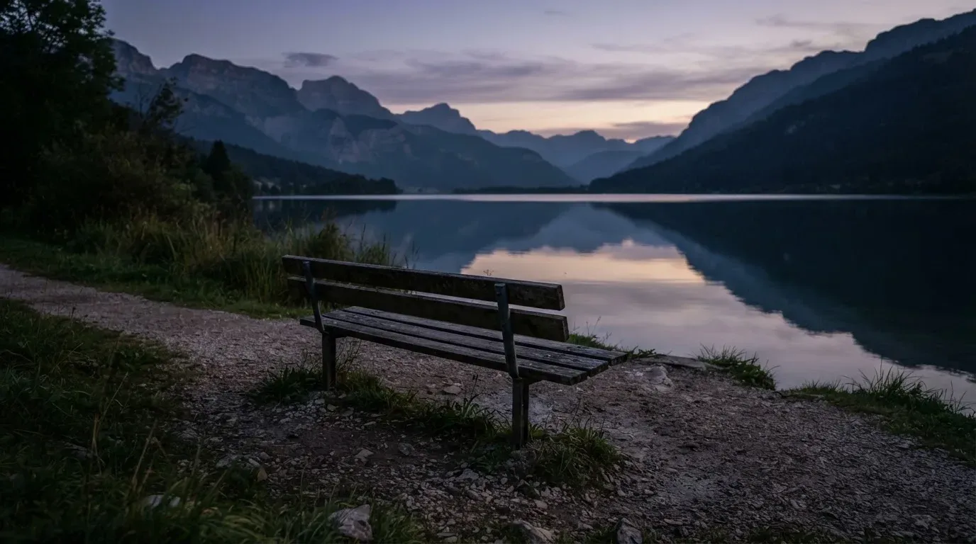 Eine leere Holzbank am Seeufer in der Dämmerung, im Hintergrund dunkle Berge