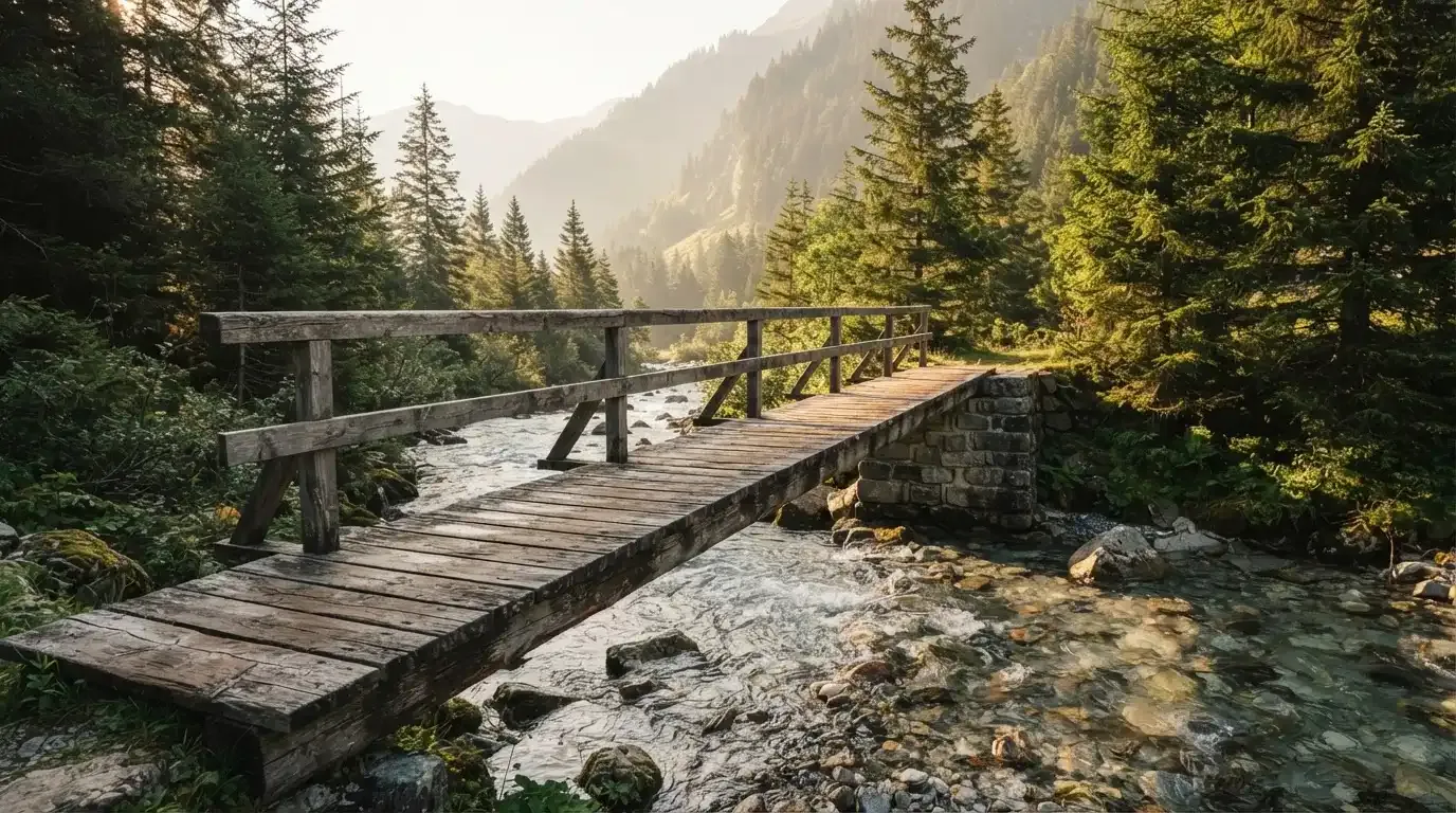 Eine alte Holzbrücke führt über einen Bergbach im Morgenlicht, Tannen säumen das Ufer