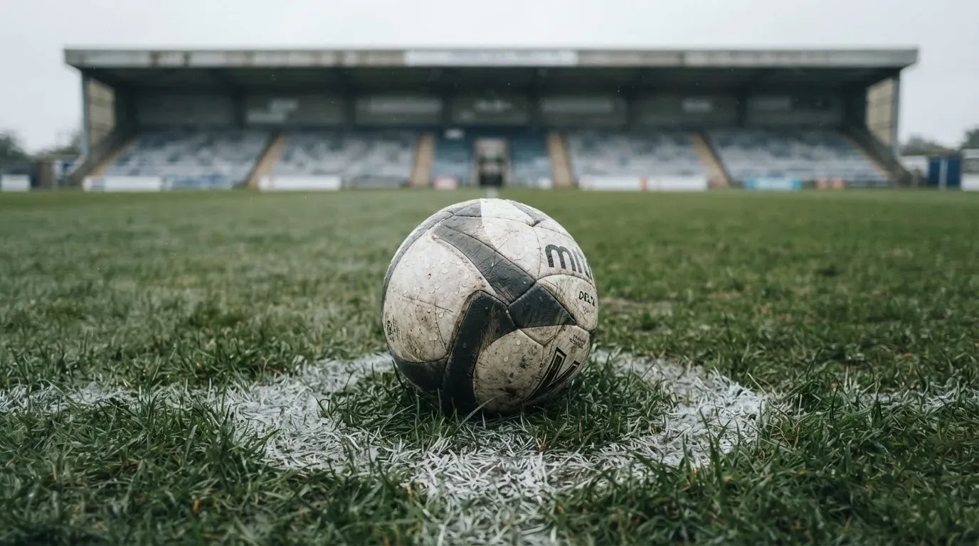Ein Fussball liegt auf dem Anstosspunkt eines nassen Rasens, dahinter eine leere Tribüne unter grauem Himmel.
