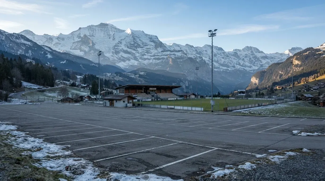 Schneebedeckte Berge ragen hinter einem kleinen Stadion in einem Alpental auf, im Vordergrund ein leerer Parkplatz am Morgen.