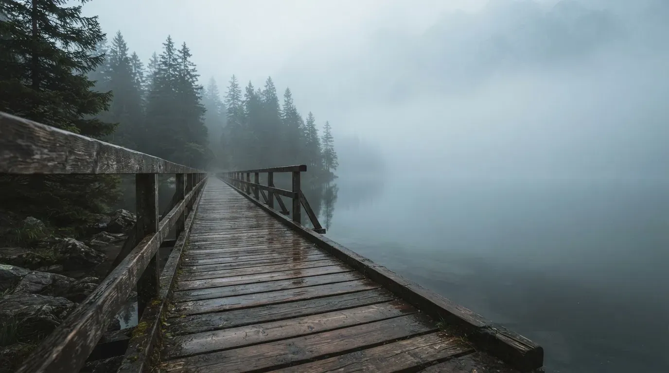 Ein Holzsteg führt über einen ruhigen Bergsee im Nebel, am anderen Ufer stehen dunkle Tannen.