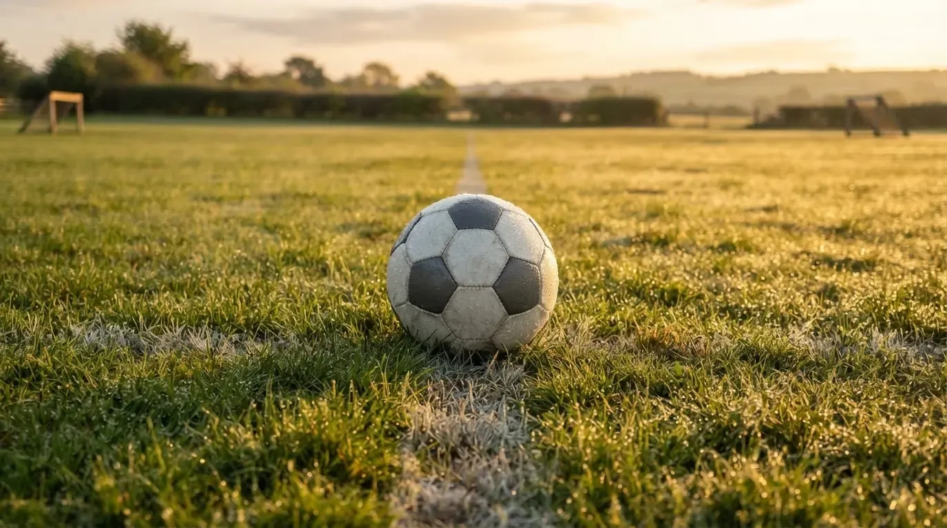 Ein Fussball liegt im Tau auf der Mittellinie eines Dorfplatzes bei Sonnenaufgang, der Rasen glitzert.