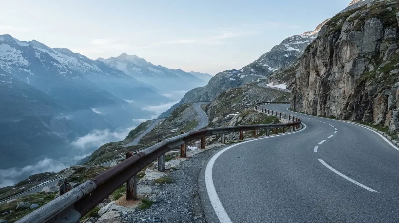 Eine Passstrasse in den Alpen mit enger Kurve und Leitplanke, darüber ein klarer Morgenhimmel und einzelne Nebelschwaden im Tal.