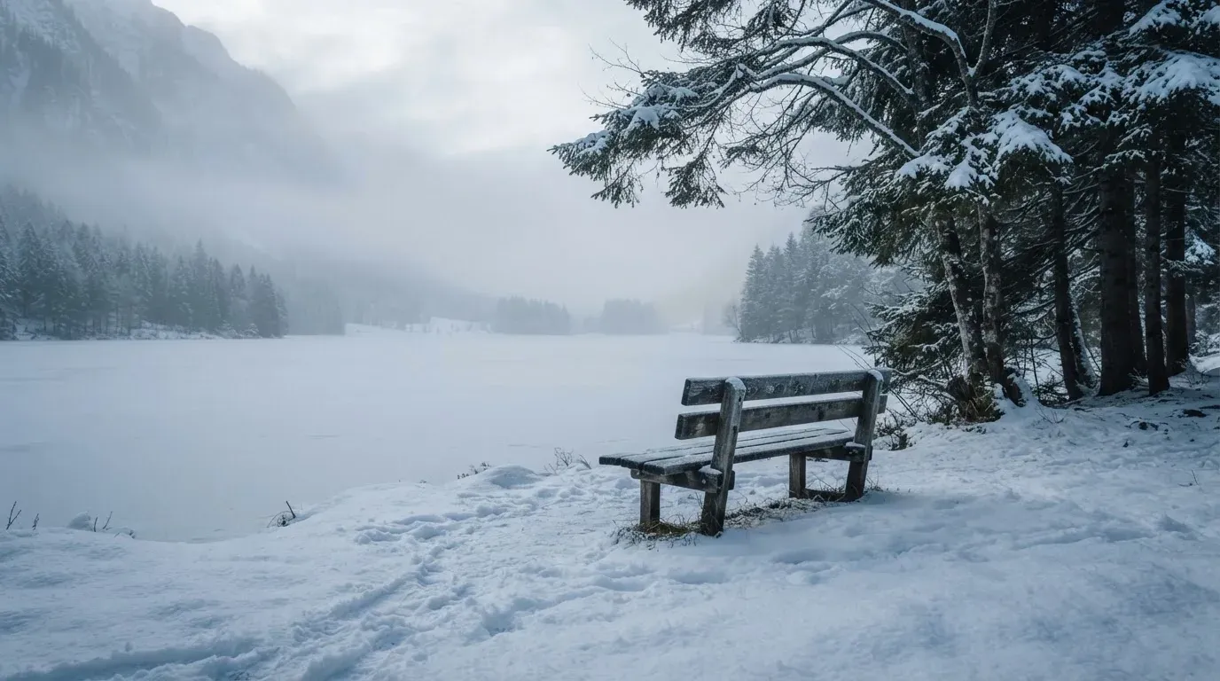 Ein zugefrorener Bergsee im Morgendunst, am Ufer steht eine einsame Holzbank zwischen verschneiten Tannen.