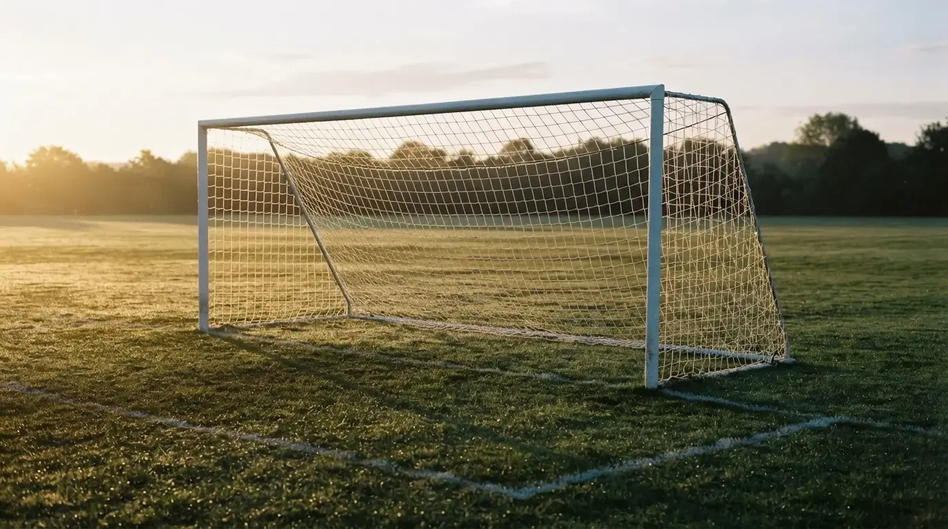 Ein Fussballtor mit Netz in der Totalen auf einem leeren Platz, Tau glänzt im Gras und die Morgensonne steht tief.