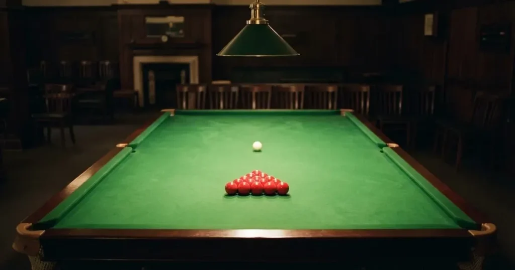 Ein Snookertisch mit grünem Tuch und einem Dreieck aus roten Kugeln unter einer tief hängenden Lampe in einer leeren Halle.