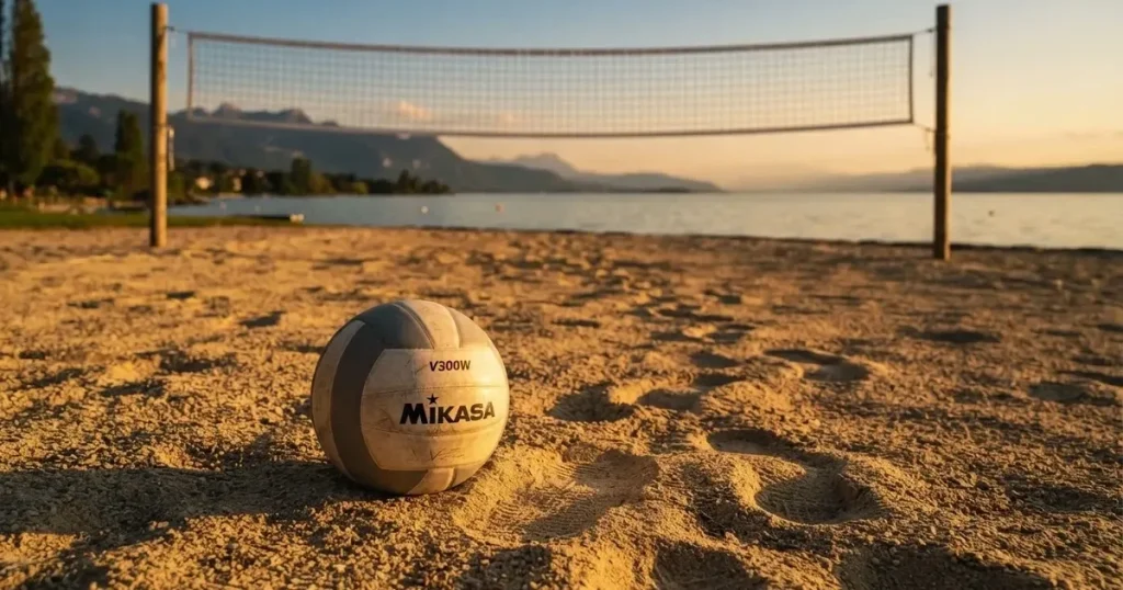 Ein Volleyball liegt im Sand eines Beachplatzes, zwischen zwei Pfosten ist ein Netz gespannt, tiefe Abendsonne wirft lange Schatten.