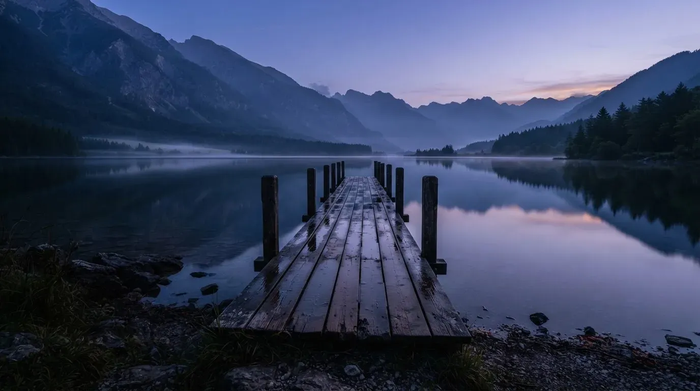 Ein Bergsee in der Dämmerung mit einem leeren Holzsteg, die Wasseroberfläche ist ruhig und Nebel zieht über die Hänge.