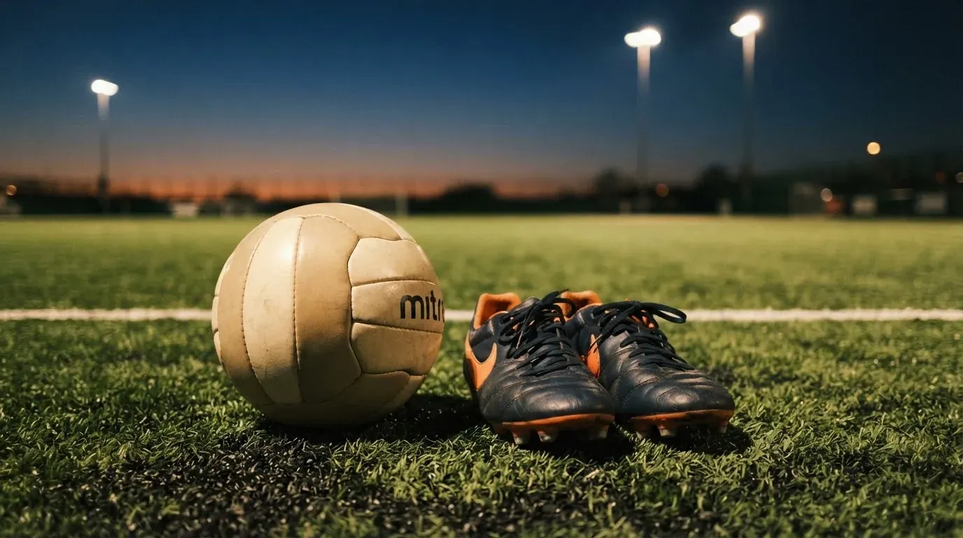 Ein Fussball und ein Paar Stollenschuhe stehen am Rand eines Kunstrasenplatzes, Flutlichtmasten ragen in den Abendhimmel.