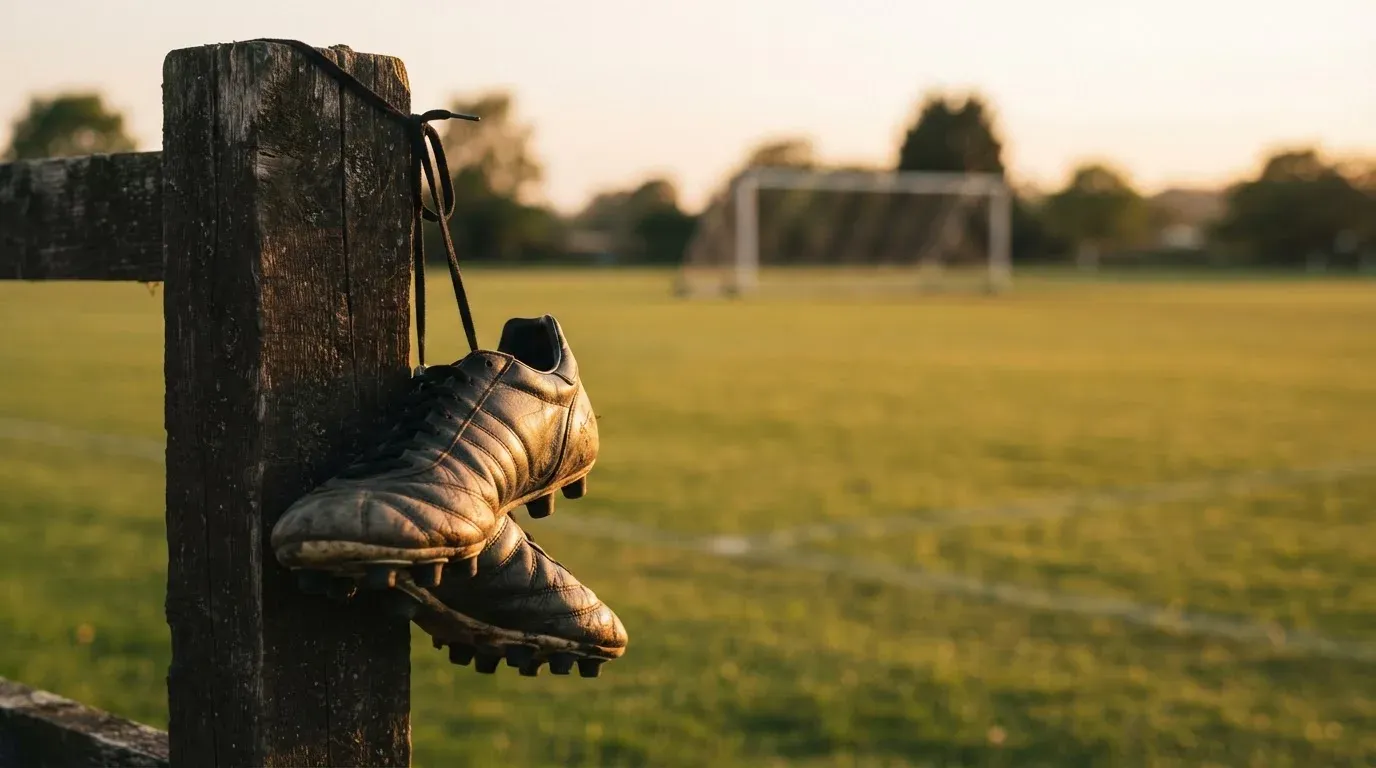 Ein Fussballschuh hängt an den Schnürsenkeln an einem Zaunpfosten neben einem Trainingsplatz im Abendlicht.