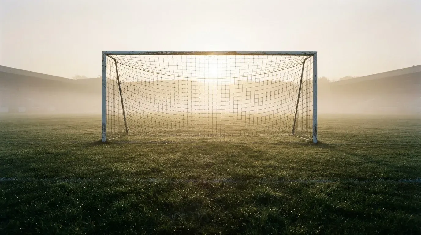 Ein Torpfosten mit Netz steht im Gegenlicht auf einem leeren Fussballplatz, über dem Rasen liegt Nebel.
