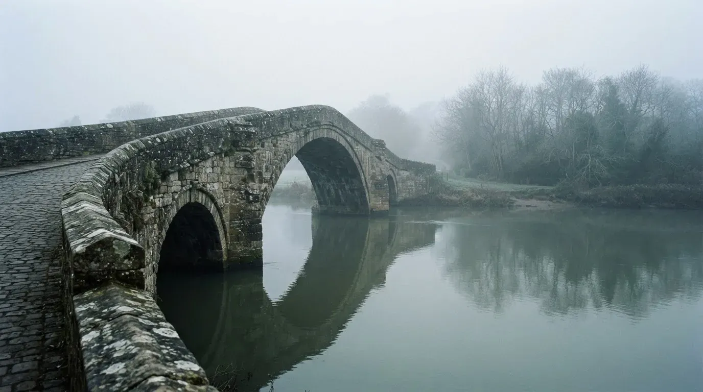 Eine Steinbrücke führt im Frühnebel über einen Fluss, auf dem Fussweg ist niemand zu sehen.