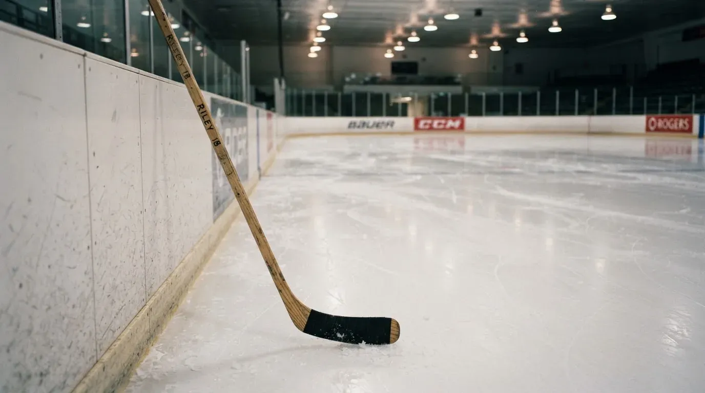 Ein Eishockeyschläger lehnt an der Bande einer leeren Halle, auf dem Eis spiegeln sich die Deckenlampen.