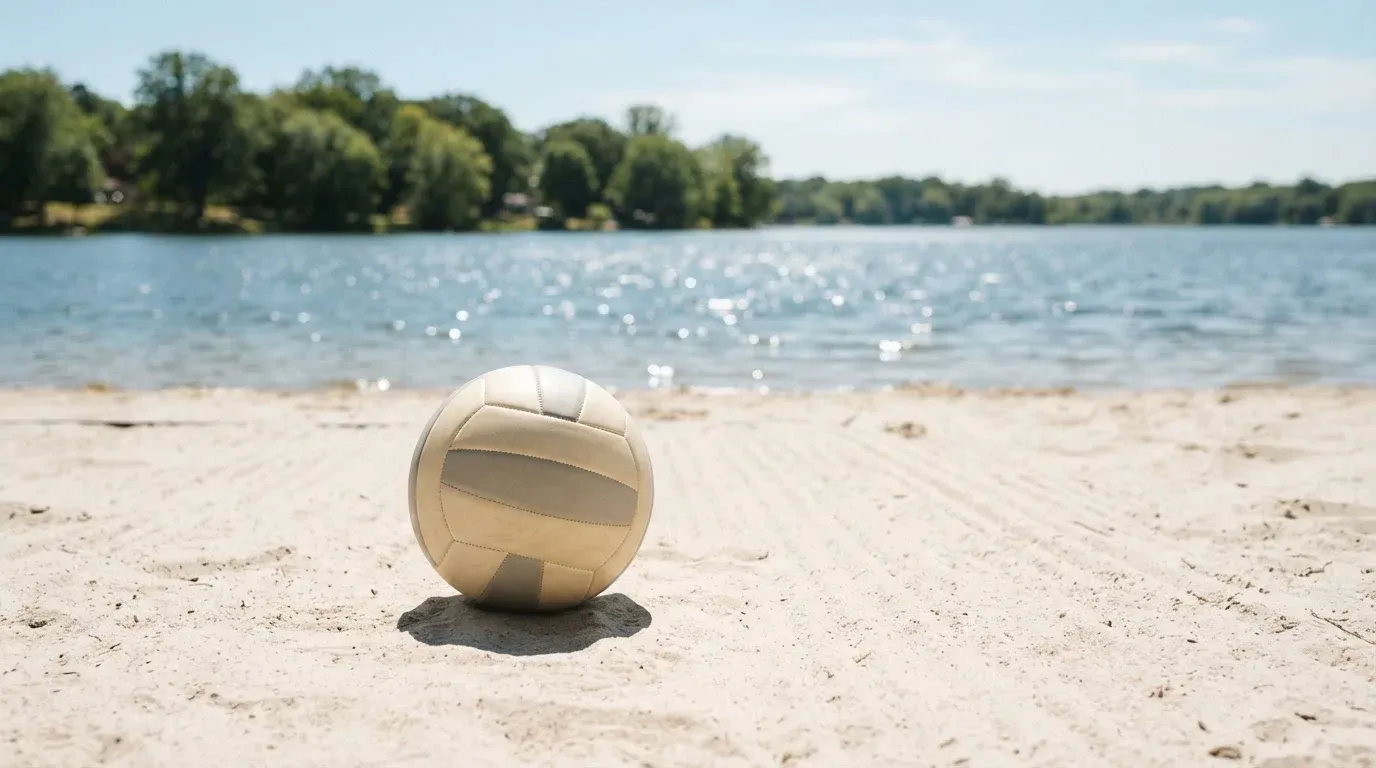 Ein Volleyball liegt auf dem Sand eines Strandfeldes am Seeufer, im Hintergrund glitzert das Wasser.