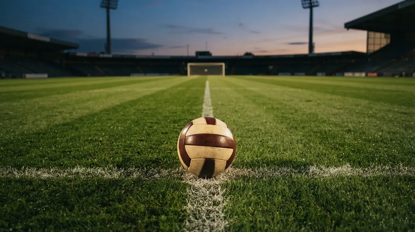 Ein Fussball liegt auf dem Mittelpunkt eines leeren Rasenplatzes, im Hintergrund ragen Flutlichtmasten in die Abenddämmerung