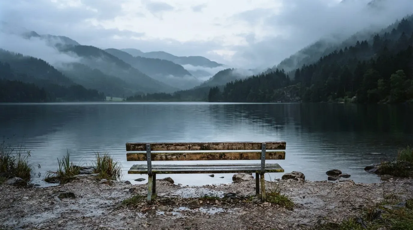 Eine Holzbank am Ufer eines ruhigen Bergsees, am gegenüberliegenden Ufer steigen dunkle Hänge in den Frühnebel