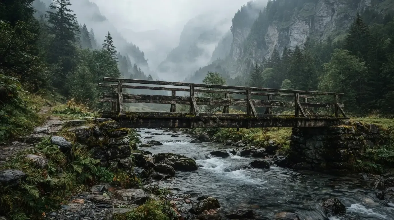 Eine alte Holzbrücke über einen Gebirgsbach, Moos auf den Steinen, Nebelschwaden hängen im Tal