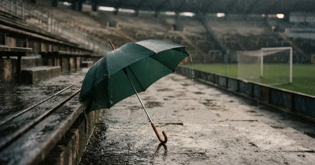 Ein offener Regenschirm lehnt an einer leeren Tribünenbank, Regen fällt auf die Stufen eines Stadions
