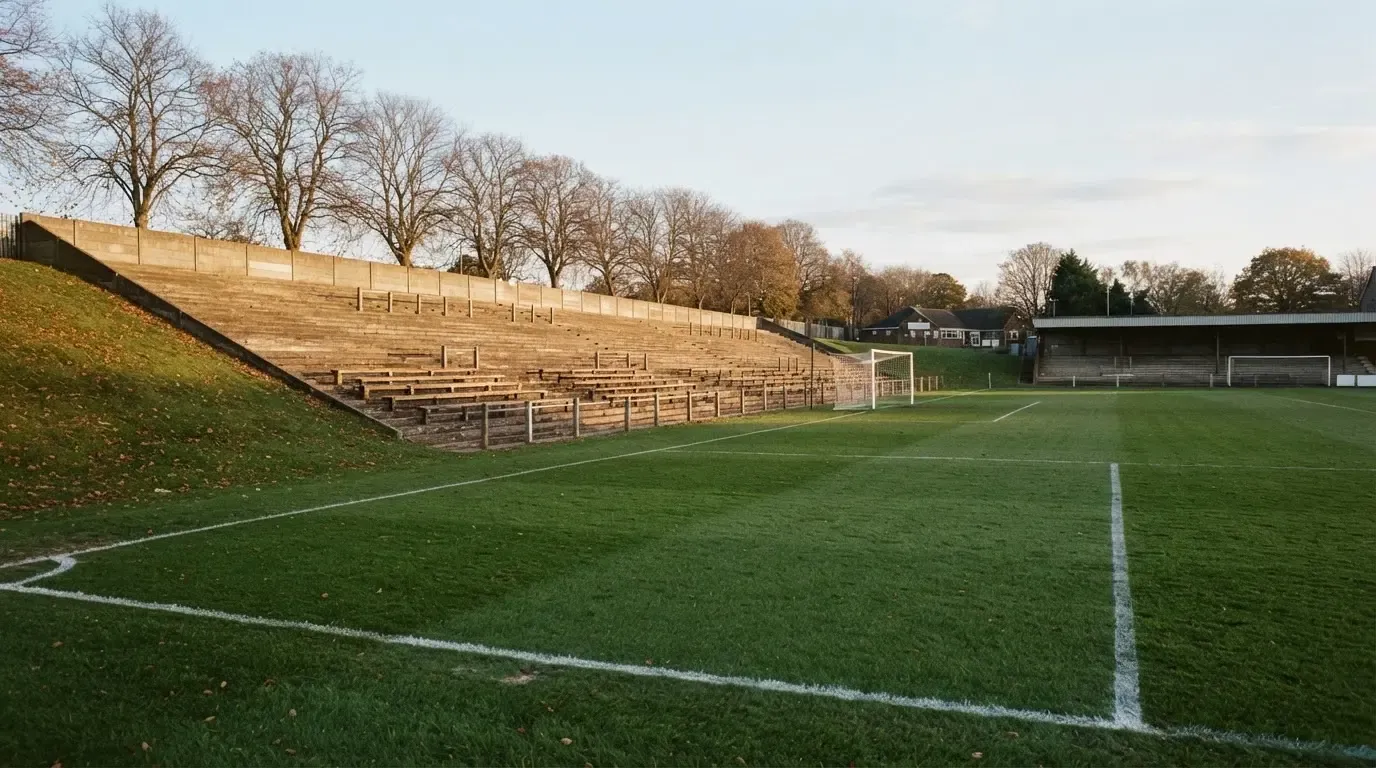 Leere Zuschauerränge eines kleinen Stadions im Herbstlicht, davor ein Rasenplatz mit frisch gezogenen Linien.