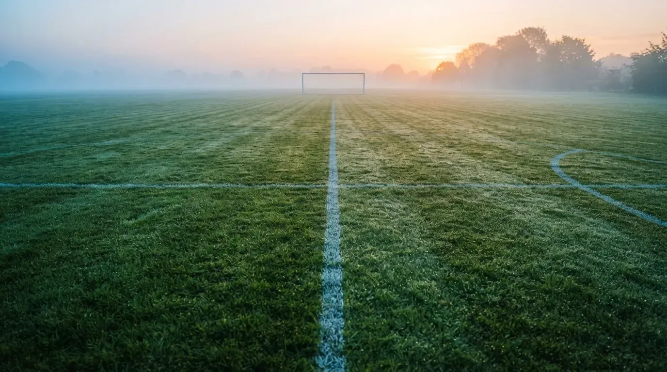 Ein Rasenspielfeld bei Sonnenaufgang, die Mittellinie verläuft quer durchs Bild und Tau glitzert auf dem Gras.