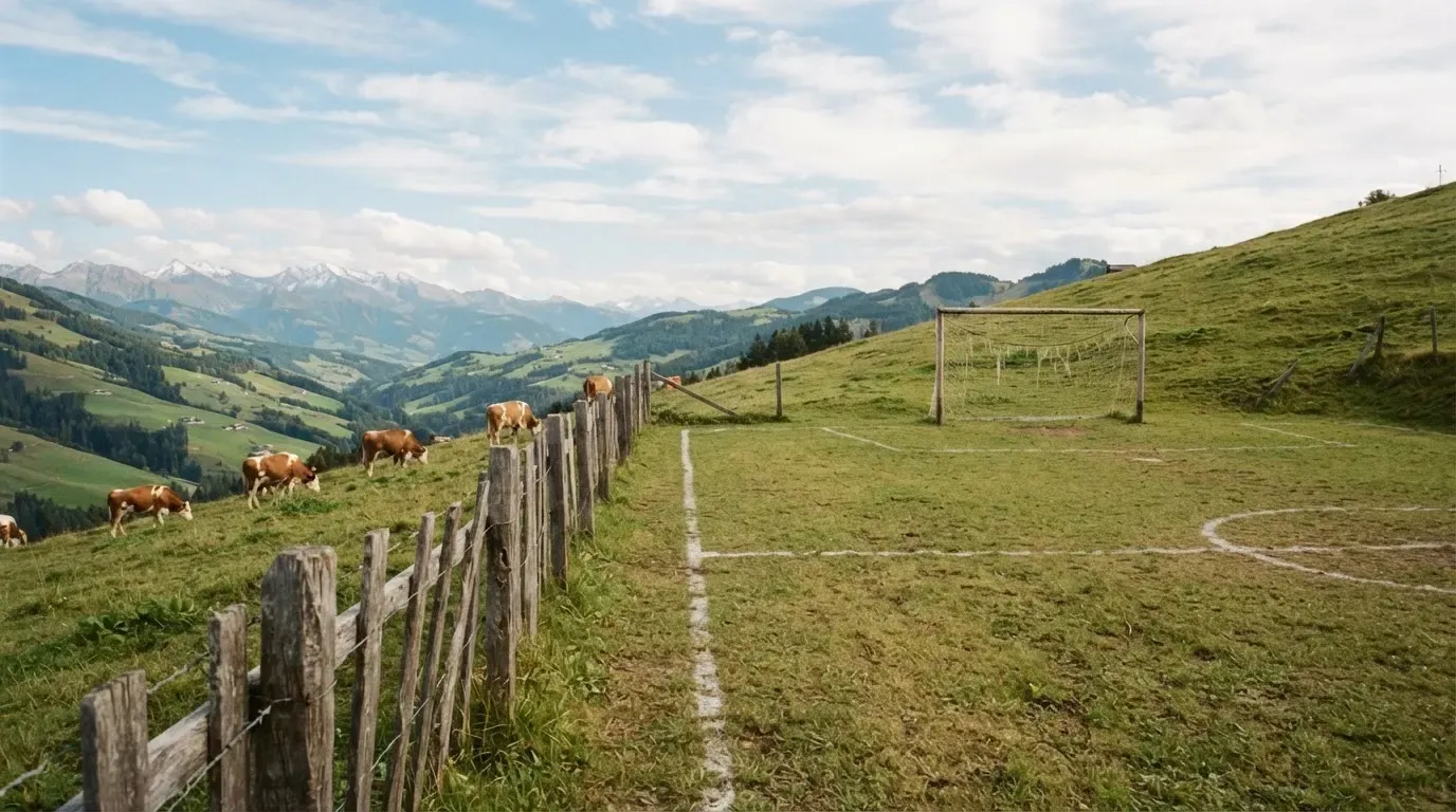 Ein Dorffussballplatz mit Holzzaun am Hang, dahinter grasen Kühe auf einer Weide unter hohen Wolken.