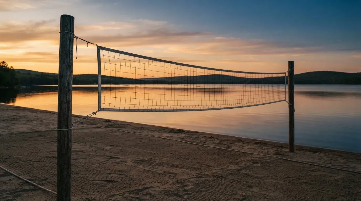 Ein Volleyballnetz steht an einem Seeufer in der Abenddämmerung, der Sand ist leer und das Wasser glatt.