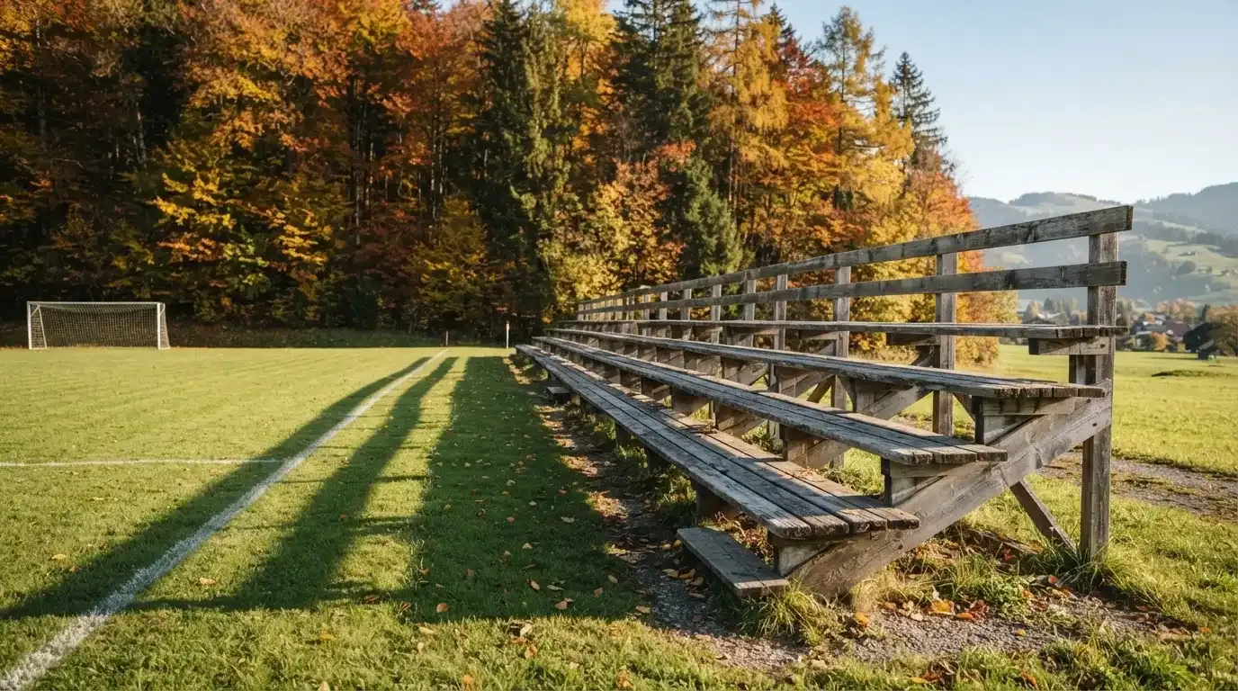 Eine leere Holztribüne steht am Waldrand neben einem kleinen Sportplatz im Herbstlicht.