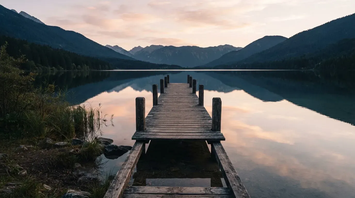 Ein leerer Holzsteg ragt am Abend in einen ruhigen Bergsee, das Wasser spiegelt den Himmel.