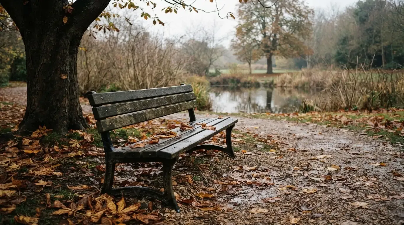 Eine Parkbank unter einer Kastanie, Herbstlaub bedeckt den Kiesweg, im Hintergrund ein stiller Teich.