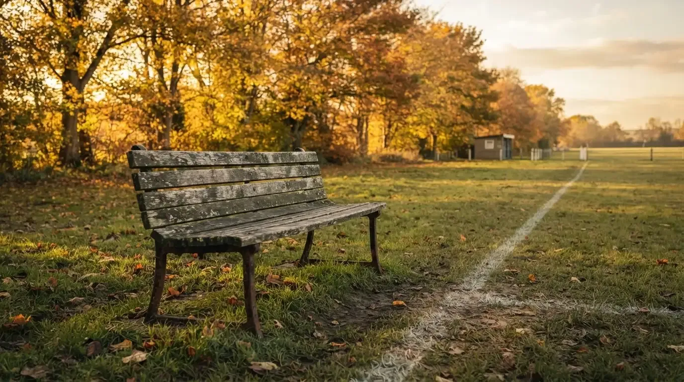 Eine hölzerne Bank steht an der Seitenlinie eines leeren Sportplatzes, dahinter Bäume im Herbstlicht.
